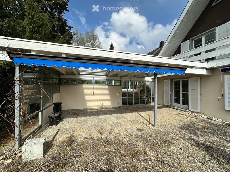 House with a covered patio, blue awning, glass walls, and a gravel floor.