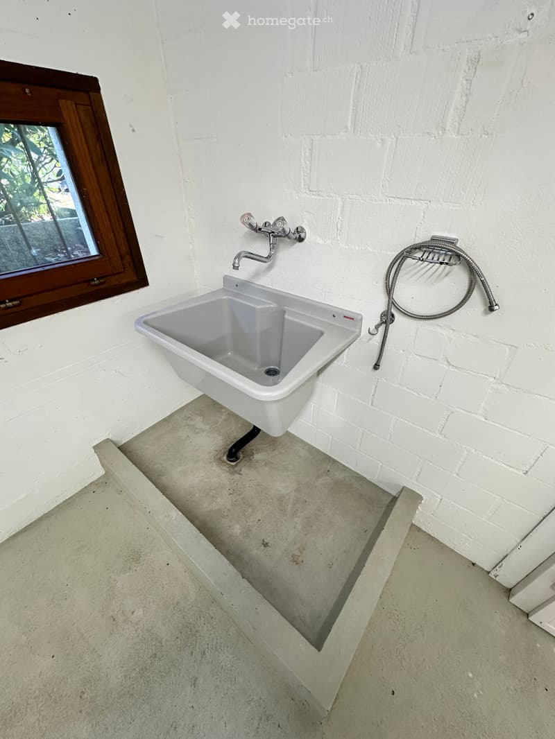 A small room with a white sink, faucet, and shower hose attached to the wall. The sink is placed on a concrete platform. The wall is made of white bricks, and there is a wooden window frame with a glass window.
