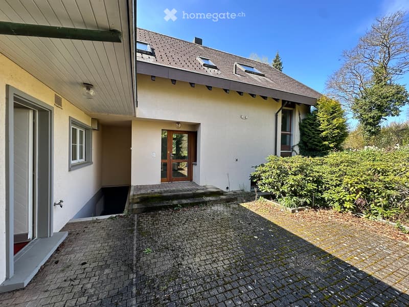 Raised ground floor, white exterior, brown roof, wooden door, brick driveway, shrubs, trees
