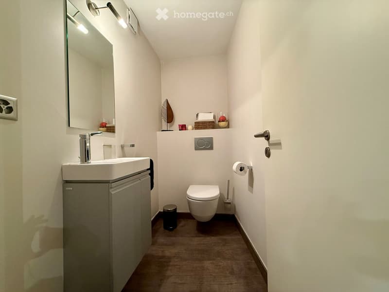 small bathroom with white walls, wooden floor, white sink, mirror, toilet, trash can, toilet paper