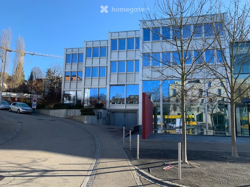 Modern office building with glass facade, multiple floors, entrance with automatic doors, parking area, street with cars parked, trees, and a crane in the distance.