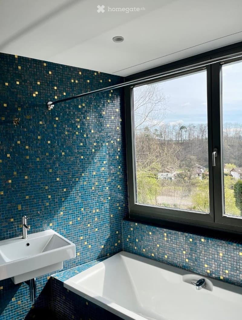 Bathroom with blue and gold mosaic tiles, white bathtub, white sink, large window with view of trees and houses
