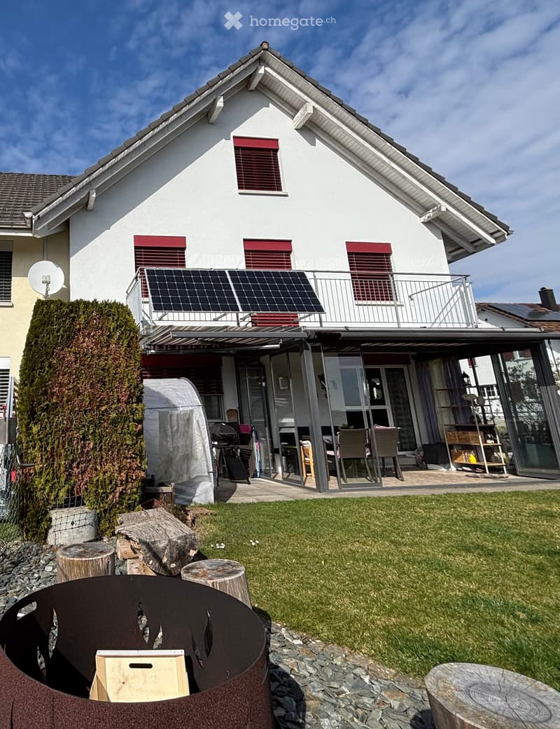 Detached house, white exterior, red trim, solar panels on the balcony, fenced garden, green lawn, seating area with chairs and table