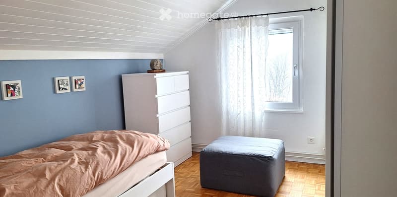 Bedroom with bed, white dresser, ottoman, three framed pictures on wall, window with white curtains, wooden floor