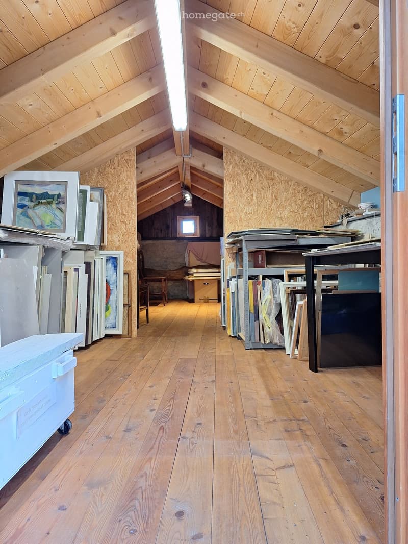 A room with wooden floors, wooden beams, and a ceiling with a long light. There are several paintings and frames on the wall, and a table with a box on it. There is a wooden chair and a small window in the room.