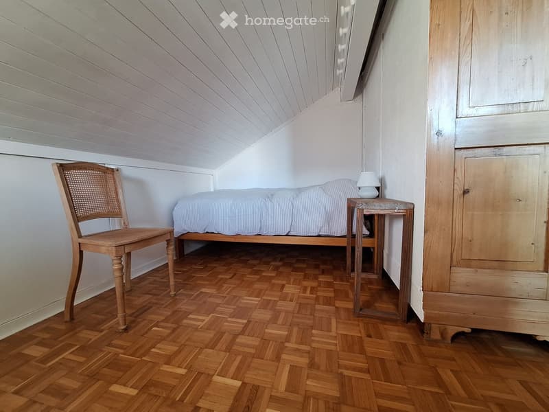 small room with wooden bed, chair, nightstand, lamp, wooden floor, white walls, wooden cabinet