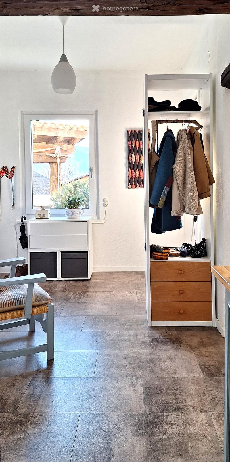 A room with white walls, wooden floor, a white cabinet with drawers, a white shelf with clothes, a window with a view of a wooden structure and plants, a hanging lamp, and a chair with a pillow.