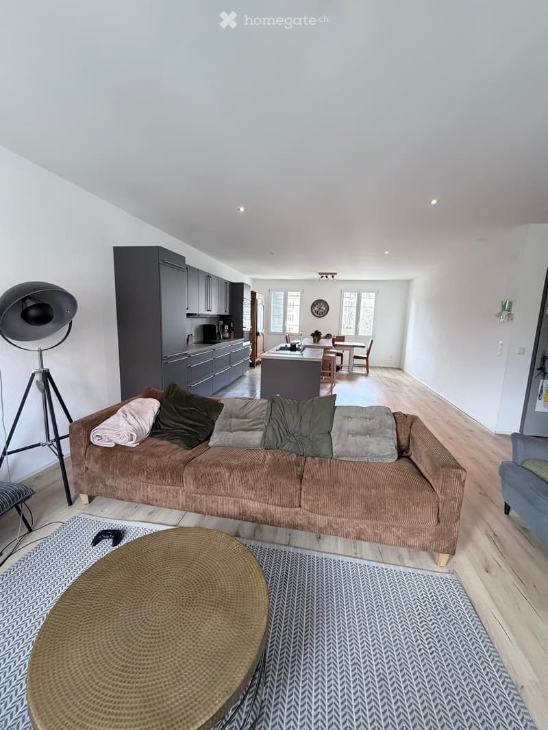 modern living room with a large brown couch, a round coffee table, a floor lamp, a rug, and a kitchen with a dining area