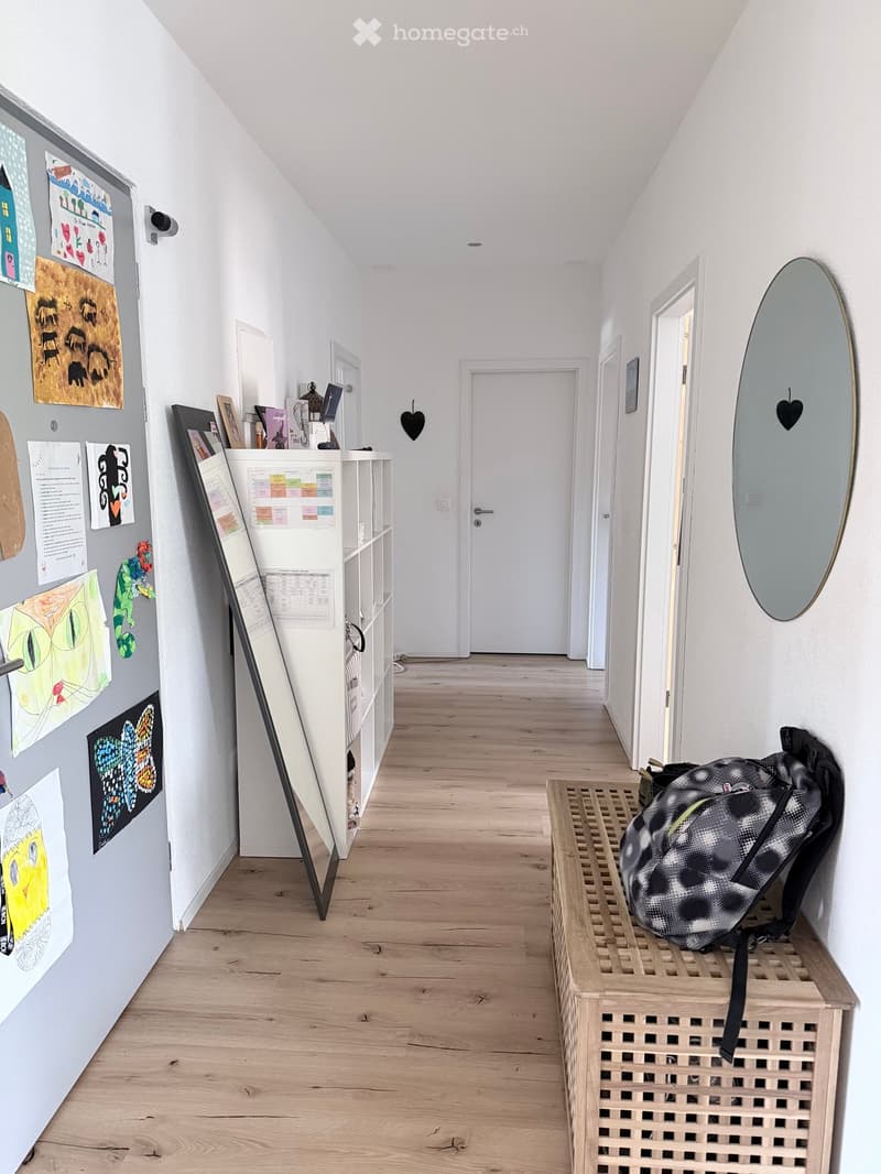 A hallway with wooden floors, white walls, a white shelf, a mirror, and a backpack on a wooden storage box.