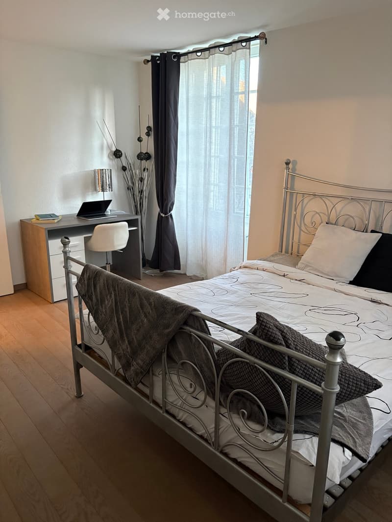 Furnished Room. A bedroom with a bed, desk, chair, and laptop. The bed has a metal frame and is covered with a blanket and pillows. The desk has a laptop, books, and a lamp. The room has a wooden floor and white walls. There is a window with white curtain