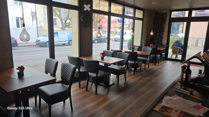 A restaurant with wooden floors, black chairs, wooden tables, large glass windows, and a counter with bottles and menus.