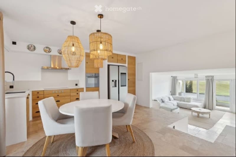 Open-plan kitchen and living room, white dining table, white chairs, wooden cabinets, white refrigerator, wooden pendant lights, white couch, glass windows, tiled floor