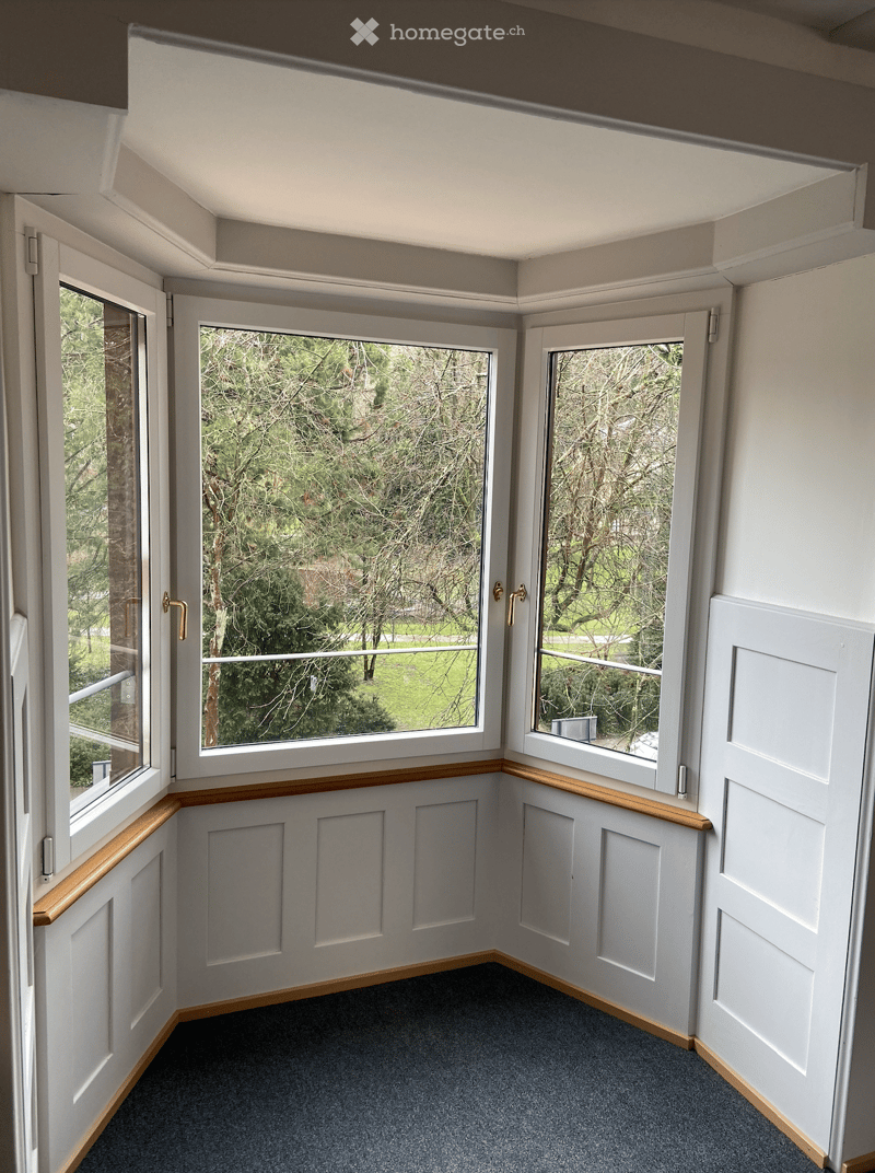 Corner room with three large windows, white walls, wooden window sills, carpeted floor, and a view of trees and grass outside.