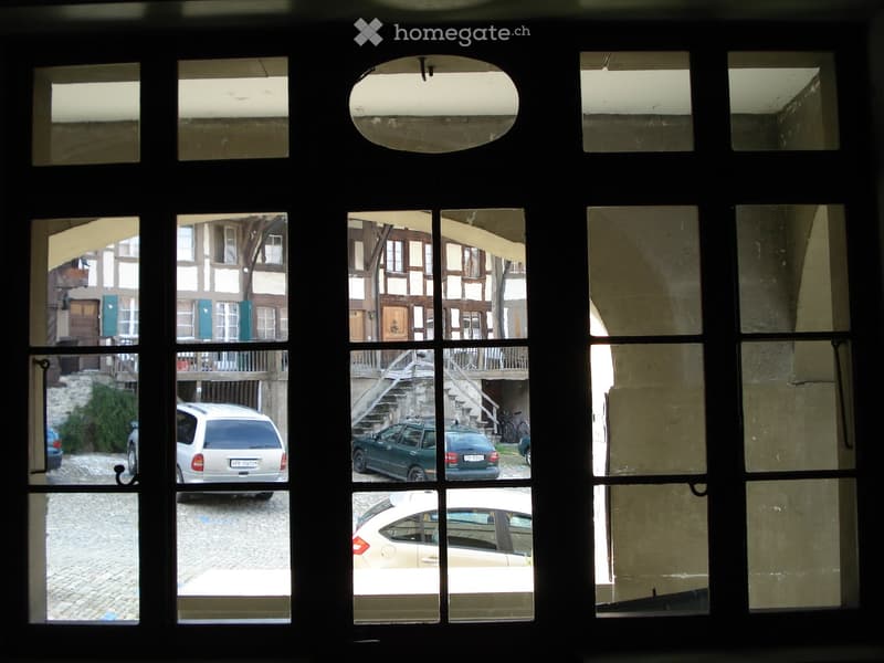 View of a parking lot with several cars parked, a building with wooden beams and stairs, and a cobblestone road.