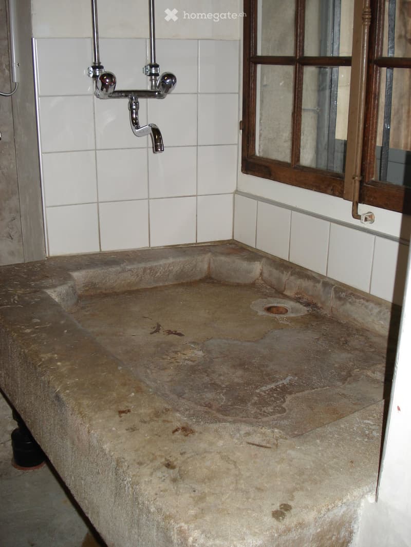 A concrete sink with a faucet mounted on the wall, white tiled walls, a wooden window frame, and a small window