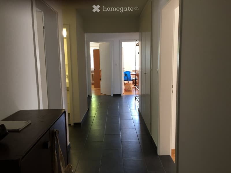 Empty hallway with tiled floor, white walls, open doors, and a wooden cabinet with a book on top.