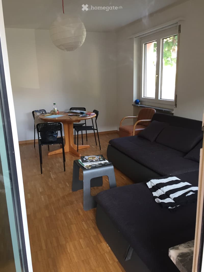 A living room with a wooden table, chairs, a couch, a side table, a lamp, and a window.