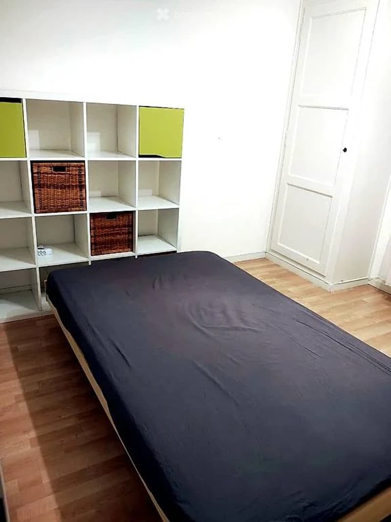 Single bed, wooden floor, white wall, white door, white shelf with green and brown baskets