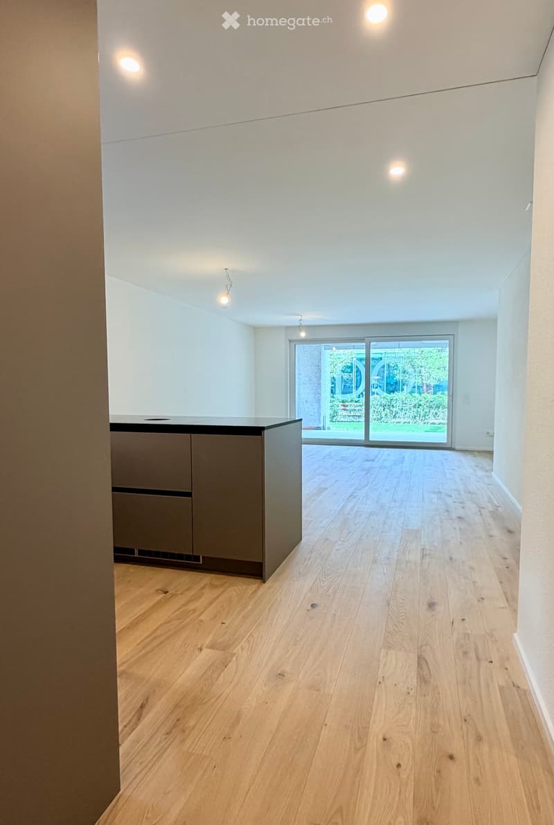 Empty room with light wooden flooring, white walls, and a small kitchen counter in the corner. Large sliding glass doors lead to an outdoor area.