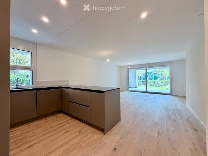 Modern kitchen with light wooden flooring, grey cabinets, and a black countertop. Features a large window and a sliding glass door leading to an outdoor area.
