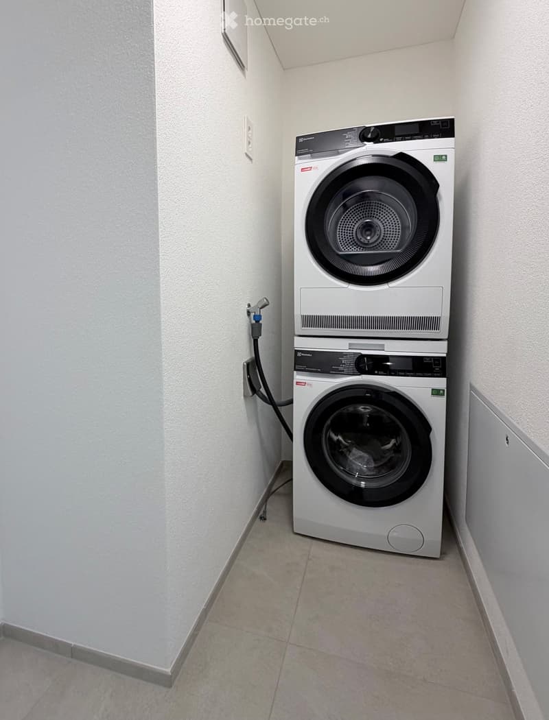 A compact bathroom with a stacked washing machine and dryer unit, white tiled walls, and a tiled floor.