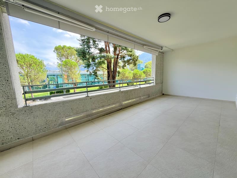 Empty balcony with tiled floor, white walls, large window offering a view of trees and mountains, metal railing, and a light fixture on the ceiling.