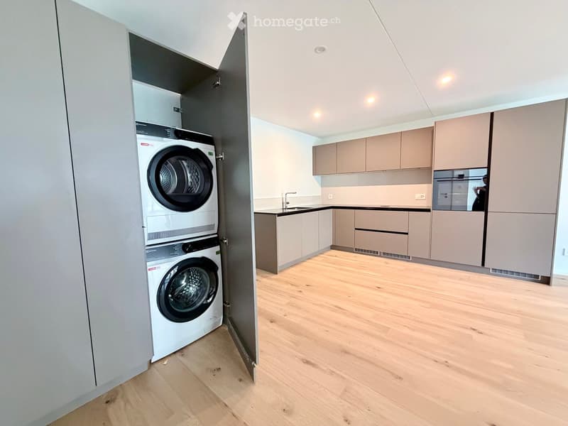 Modern kitchen with light wood flooring, light-colored cabinets, and a built-in dishwasher. A private washing machine is visible in an adjacent closet.
