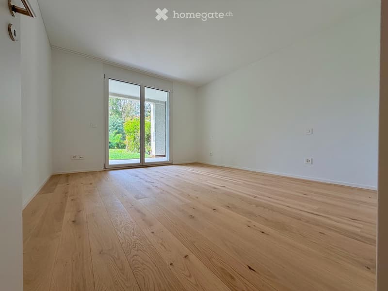 Empty room with light wooden flooring, white walls, and large sliding glass doors leading to a garden.