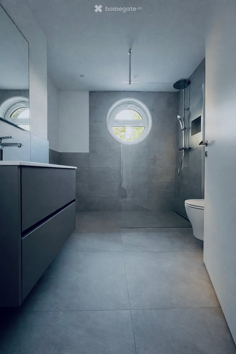 modern bathroom with gray tiles, round window, glass shower enclosure, white toilet, gray vanity with drawers, silver faucet