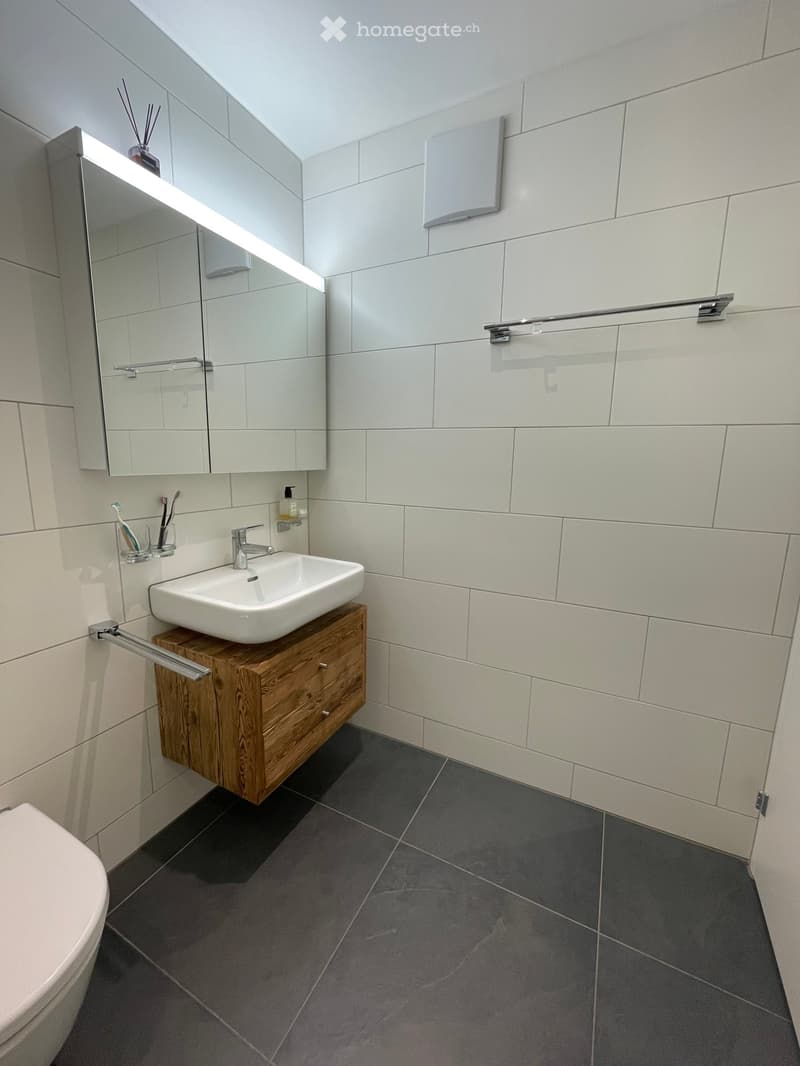modern bathroom with white tiles, wooden cabinet, white sink, mirror, toilet, and towel rack