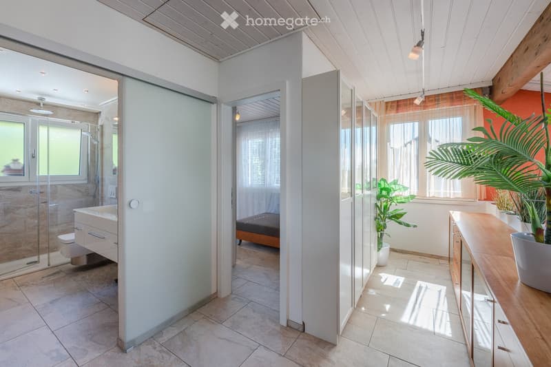 Bathroom with shower, toilet, sink, and a small bed in the corner. The room has a glass door, tiled floor, and a window with curtains. There is a wooden cabinet with a potted plant on top.