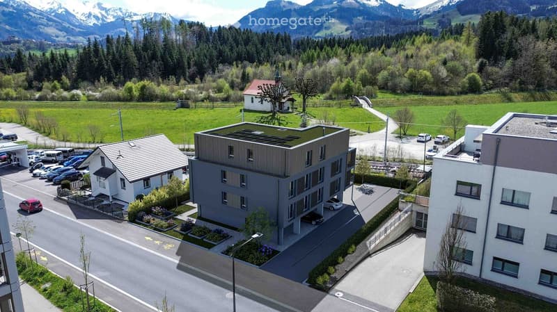 Aerial view of a modern apartment building with a parking lot, surrounded by greenery and mountains