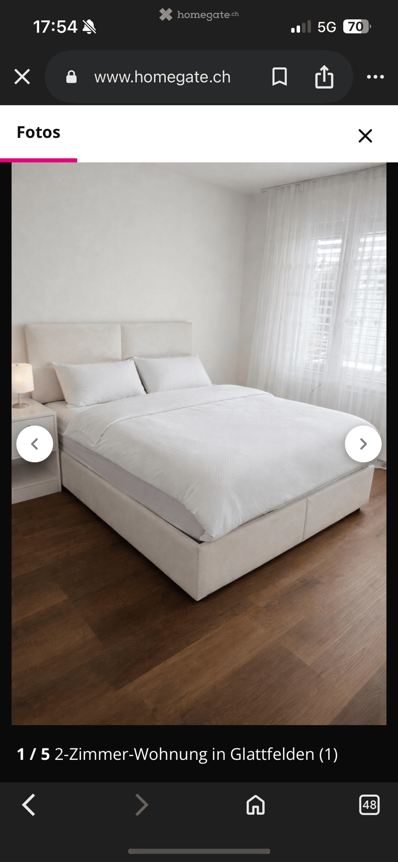 Bedroom with a bed, white headboard, white bedding, wooden floor, white curtains, nightstand with lamp, balcony