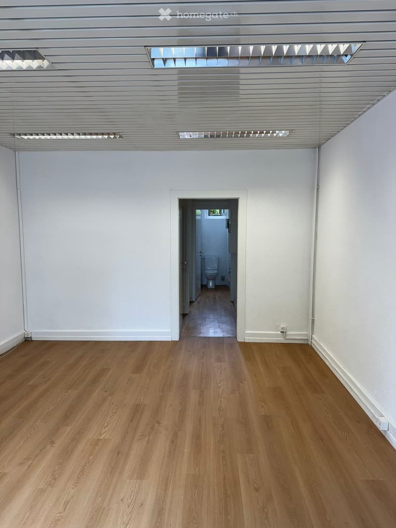An empty room with white walls, wooden floors, and a ceiling with recessed lighting. There is a door leading to a small hallway with a toilet.