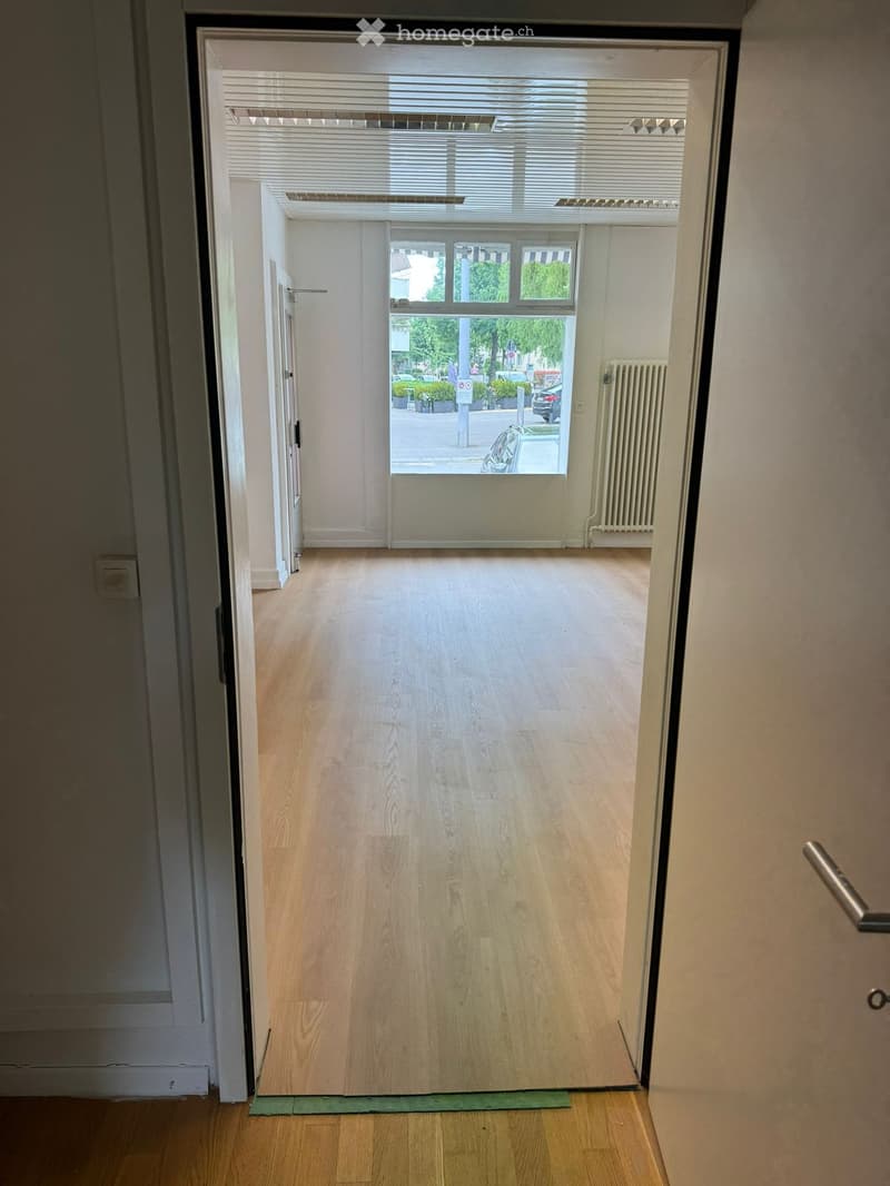 An empty room with wooden floors and white walls. The room has a large window with a view of a parking lot and trees outside. There is a white radiator on the wall and a door leading to another room.
