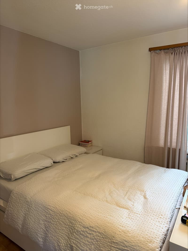 A bedroom with a bed, two pillows, a nightstand with books, and a curtained window.