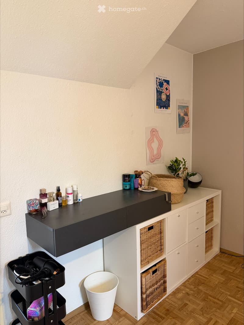 A room with a desk, shelves, and baskets. The desk has a black top and a white base. There are several items on the desk, including bottles, a basket, and a plant. The shelves are white and have baskets on them. There is a white trash can next to the desk