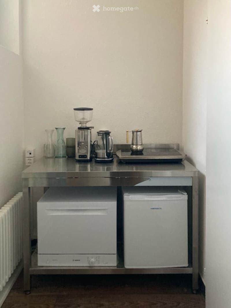 A small kitchen area with a stainless steel table, a coffee machine, a kettle, a toaster, a glass tray, a glass, a vase, and a power outlet. Below the table are a dishwasher and a washing machine.