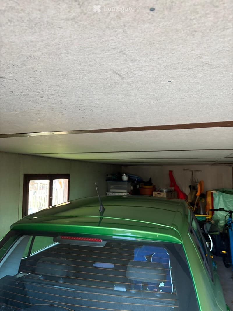 A green car parked in a garage with a white ceiling, a window, and various items stored on the shelves