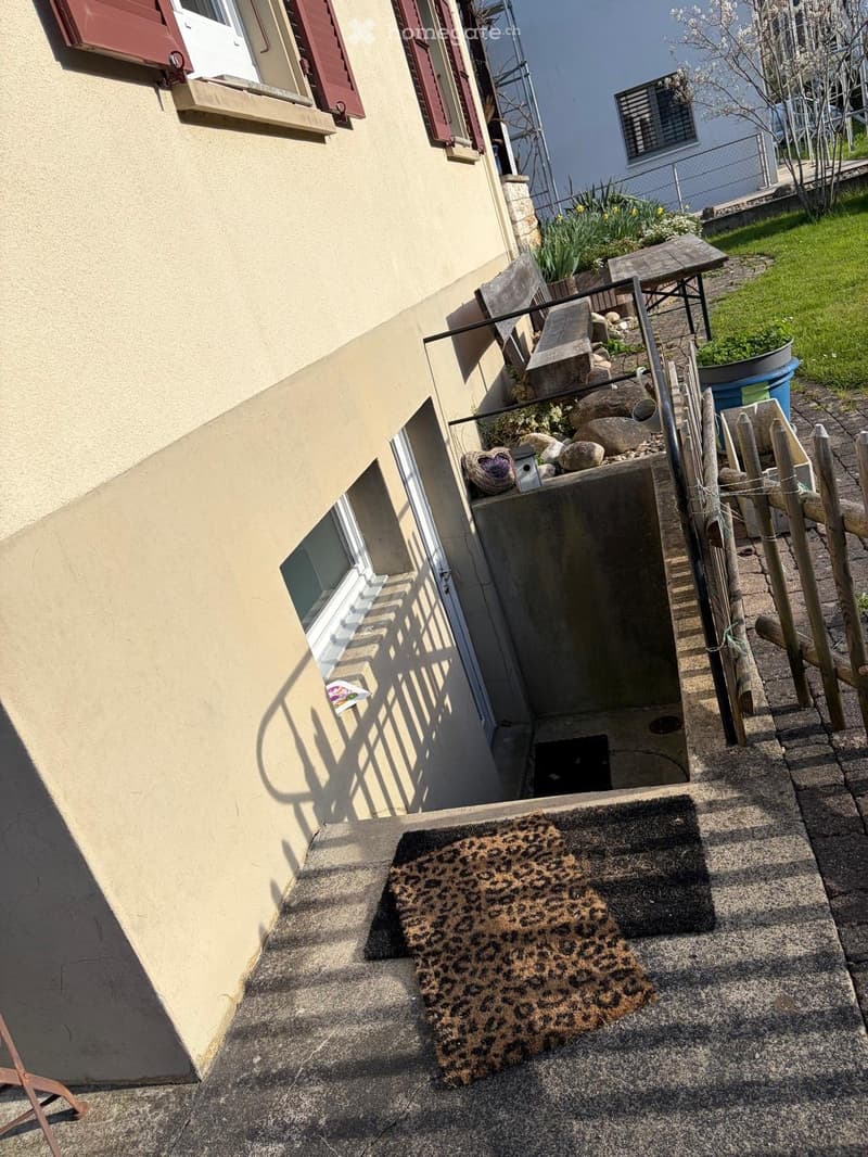 Raised ground floor, beige wall, red shutters, small window, concrete stairs, wooden bench, wooden fence, plants, grass