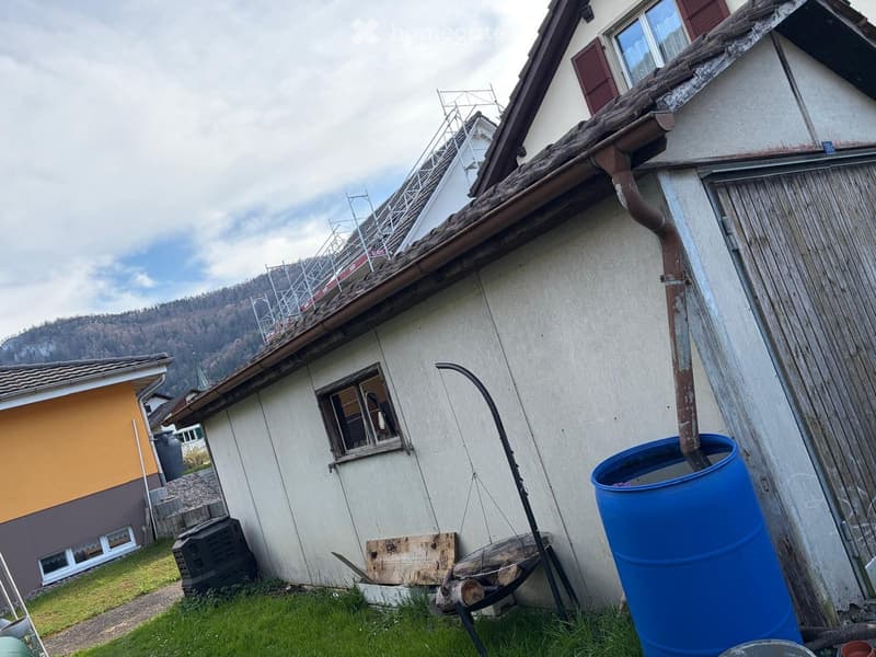 Detached house, white walls, brown roof, wooden door, window, metal scaffolding on the roof, blue barrel, metal stand, wooden plank, black object, pile of rocks, grassy area, mountain in the distance