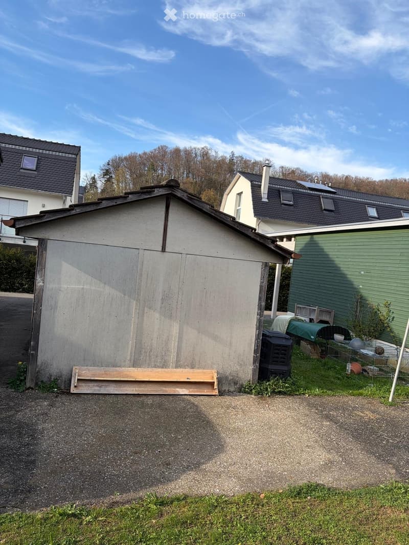 Garage in front of a house, with a wooden plank on the ground, a trash bin, and a small garden with plants and a fence.