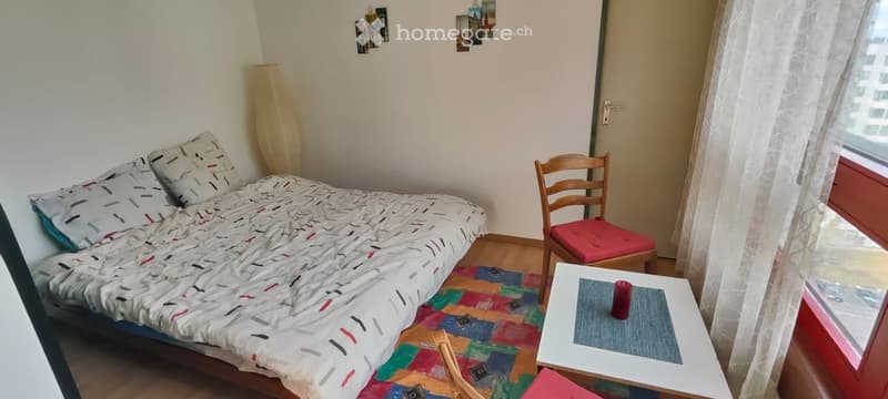 Bedroom with bed, two pillows, two chairs, table, lamp, rug, and window with curtains