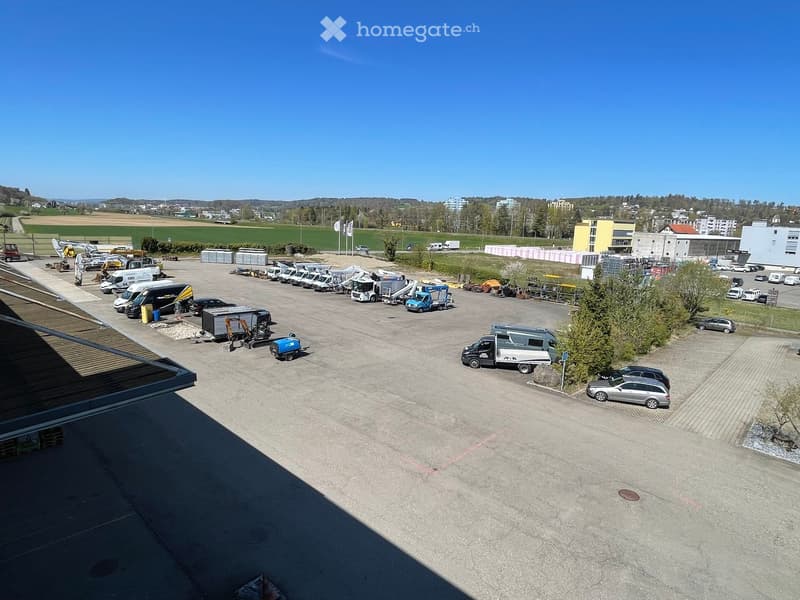 Aerial view of a parking lot with multiple vehicles parked, including trucks, vans, and cars. The parking lot is surrounded by trees and has a clear view of the surrounding area.