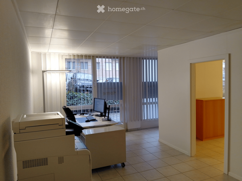 An empty office with a desk, computer, printer, and a cabinet. The room has white walls and tiled flooring. There is a window with blinds and a view of a car parked outside.