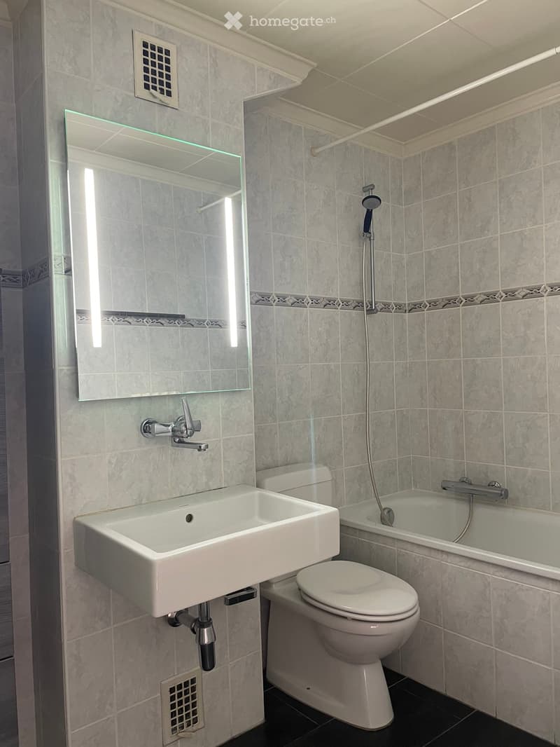 Modern bathroom with a white sink, toilet, bathtub, and large mirror with built-in lighting. Grey tiled walls and black tiled floor.