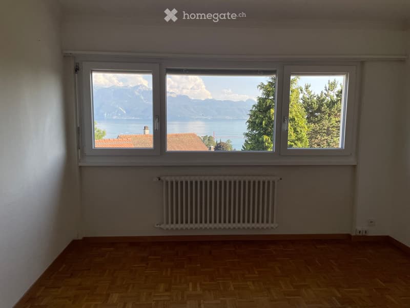 Empty room with wooden flooring, white walls, and large windows offering a scenic view of mountains and a lake.
