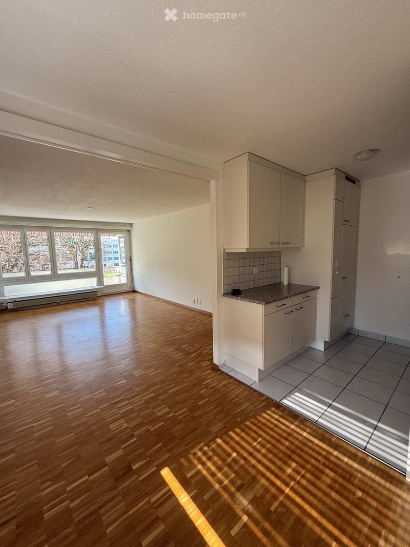 Empty room with wooden floor, white walls, large windows, kitchen area with white cabinets, granite countertop, and tiled floor.
