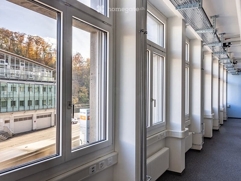 Empty room with large windows, white walls, white radiators, power outlets, and a view of a building and road outside
