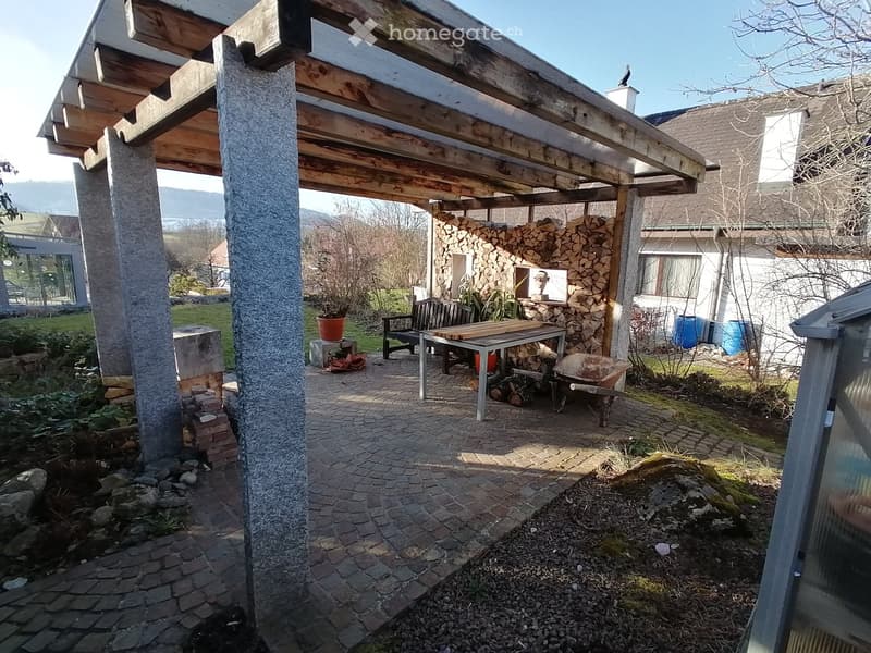 covered patio with wooden beams, brick floor, wooden table, wooden bench, potted plant, wheelbarrow, storage boxes, logs